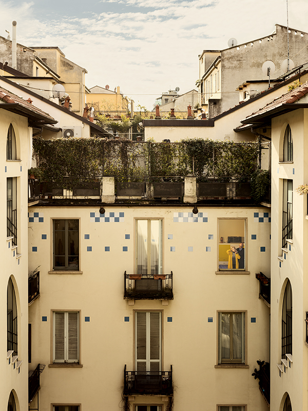 A yellow apartment building, with an elderly couple in one window. Gail Albert Halaban