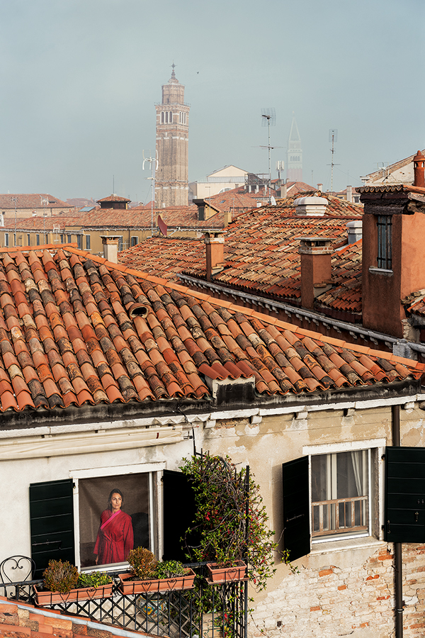 Tiled roofs of buildings beneath a blue sky. In the bottom left corner, a woman wearing a red robe looks out the window. Gail Albert Halaban