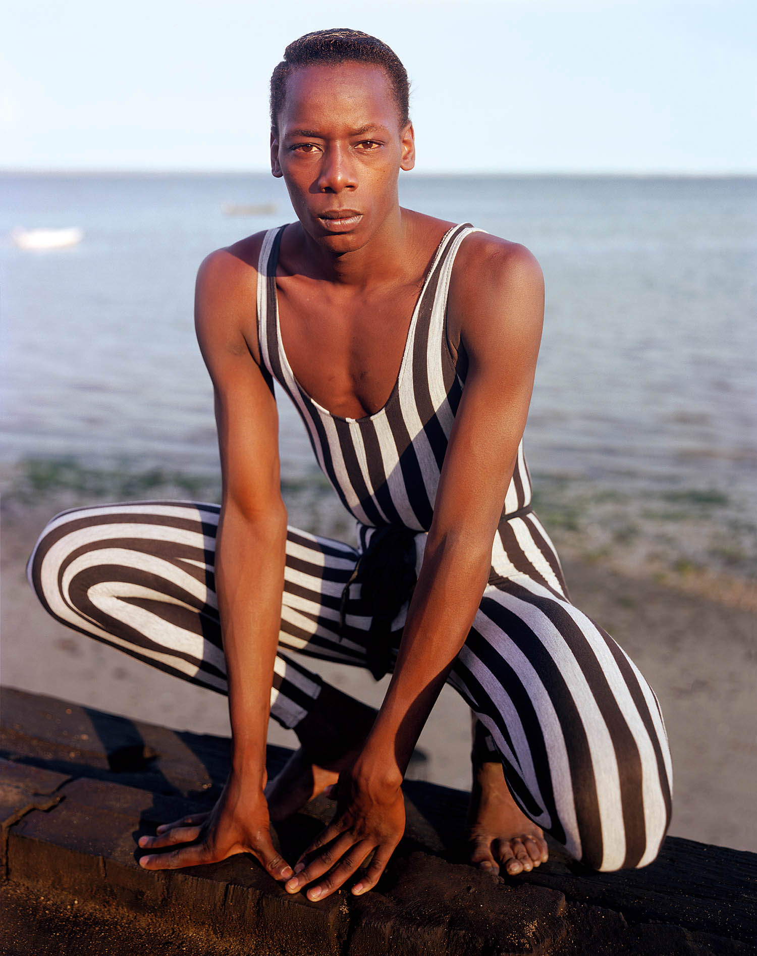 subject wearing striped bodysuit crouching on the sand in front of the ocean