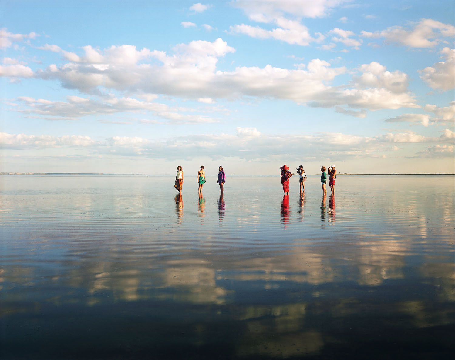 figures stand in shallow water, with the sky reflecting on the water