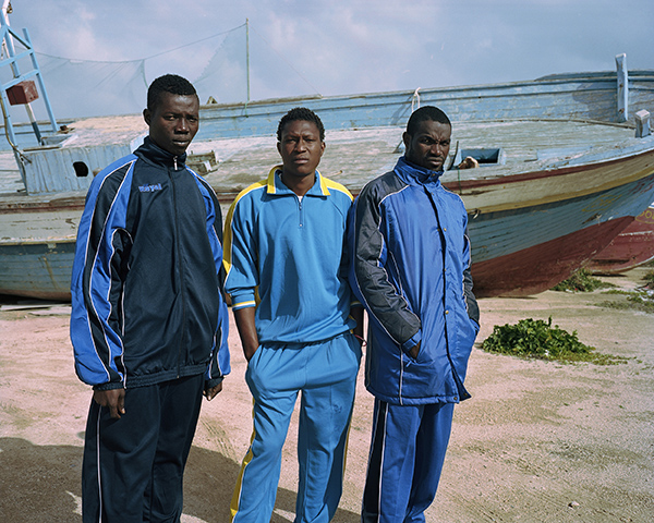 Somaro, Bouba and Abdoul

Lampedusa, Sicily, Italy, January 2017

Somaro, 19, Bouba, 19 and Abdoul, 22, arrived on the island of Lampedusa one week prior to this image being taken. The photograph was taken by the Port of Lampedusa where several migrant boats are kept as a memorial to those that crossed the Mediterranean Sea into Europe. These men experienced a particularly rough journey and all expressed the trauma of what was faced at sea. Usually vessels leave Libya in the “peek season” between May and September when seas are calmer but in this case the traffickers forced them to take the journey. They had little information regarding their situation in Lampedusa and did not know when they would be transferred to mainland Italy.
This image was taken on my second visit to the island, the first being in May 2015. On my first visit there were no migrants on the island as vessels were being diverted to Malta, Sicily and mainland Italy. Whilst driving across the town I witnessed them standing by the boats and believed it would be a suitable opportunity to photograph them and create an image that could respectfully comemorate thier journies. I was particularly drawn to their clothing and the connotations of uniform they implicate.
As can be seen in the image, the style is very simple and direct, no direction was given to the individuals other than asking them to simply stand as they wished.