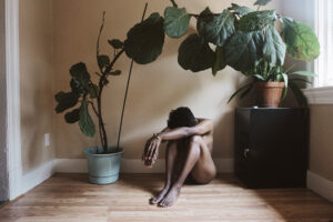 A photograph of a naked individual sitting, hiding their face, framed by a large indoor potted plant.