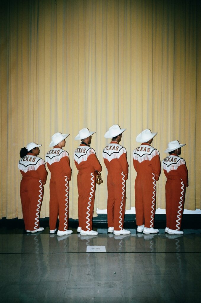 A photograph of six black marching band members in white cowboy hats facing away from the camera.The black of their matching uniforms reads "Texas."