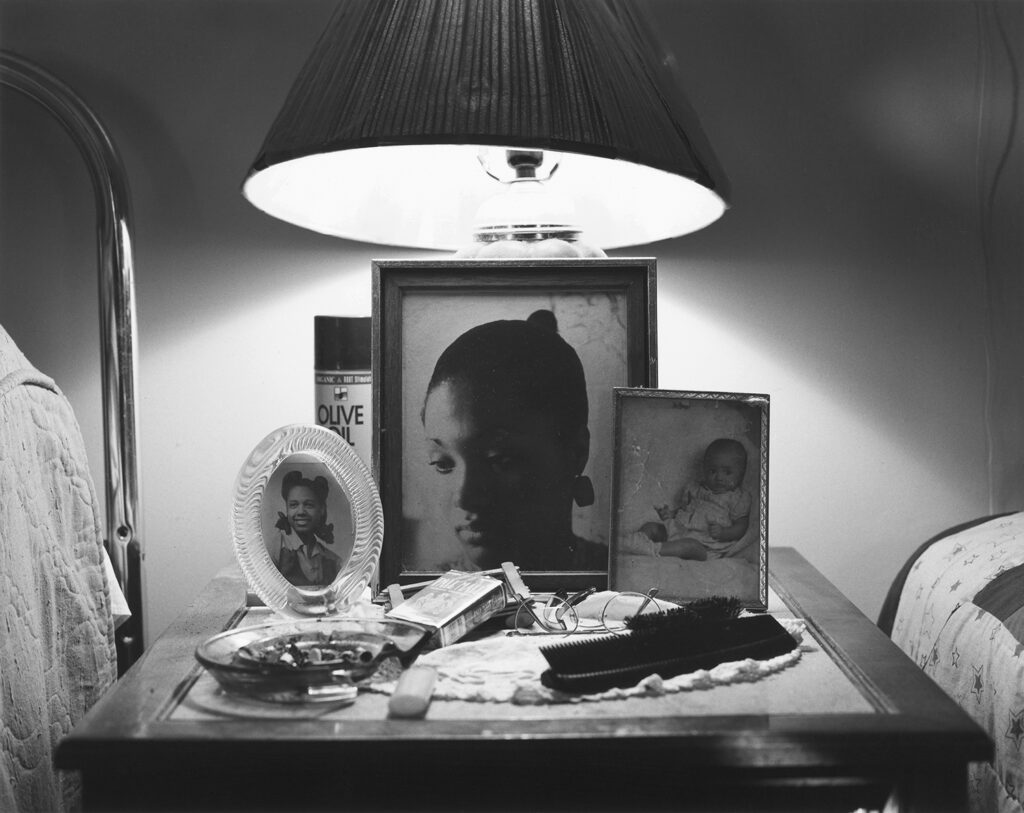 A black and white photograph of a bedside table with a lamp, framed photos, and various personal belongings.