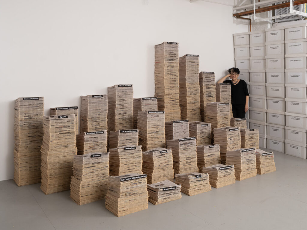 A photo of a person standing in the corner of a studio full of sculptures taking the form of stacks of papers.