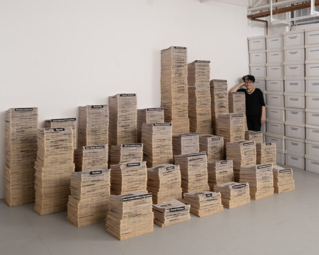 A photo of a person standing in the corner of a studio full of sculptures taking the form of stacks of papers.