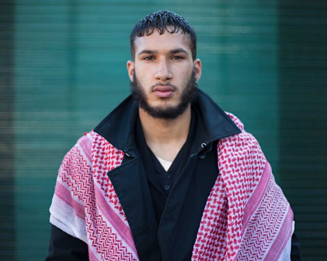 A color photograph of a Muslim man in a black coat and red scarf