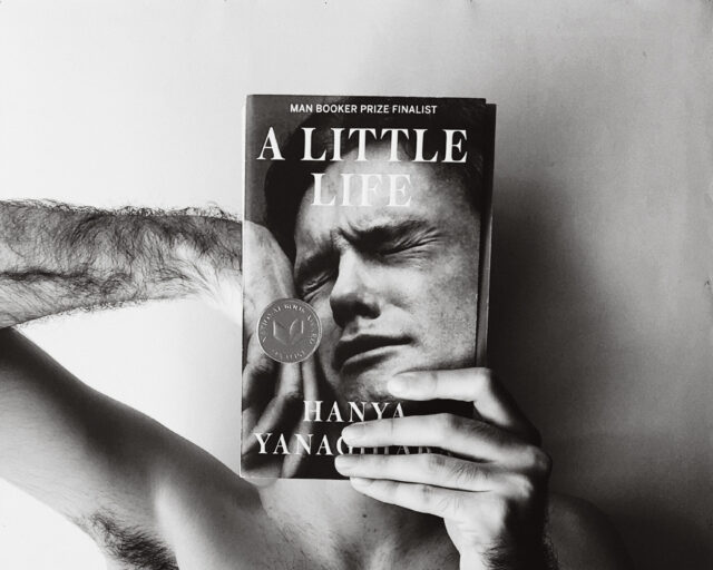 A black and white photograph of a shirtless man holding a copy of the novel A Little Life over his face