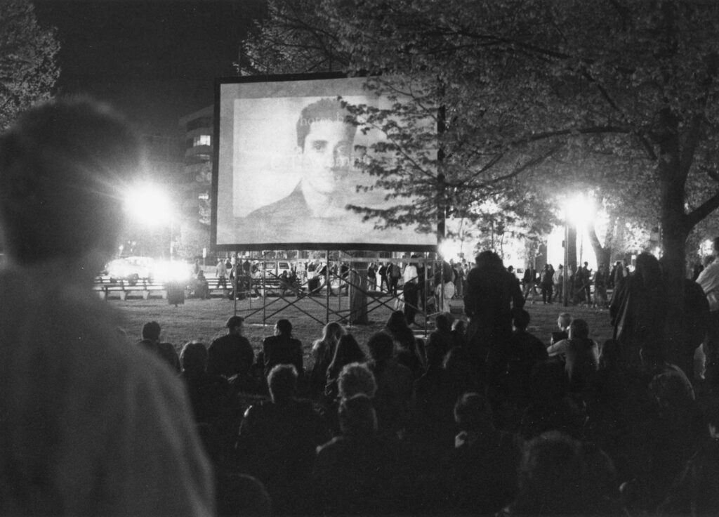 A black-and-white photograph of a crowd in a park watching a projection