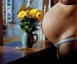 Color photograph of a pregnant white woman standing near a window with flowers