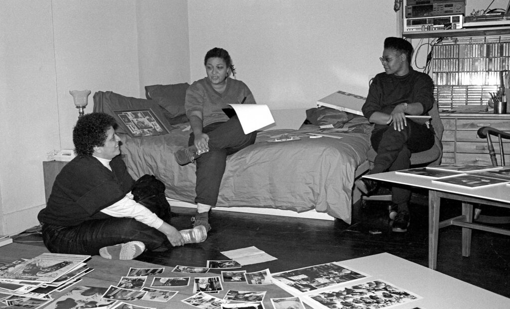 A black-and-white photo of three women in a bedroom looking at prints for a photo exhibition.