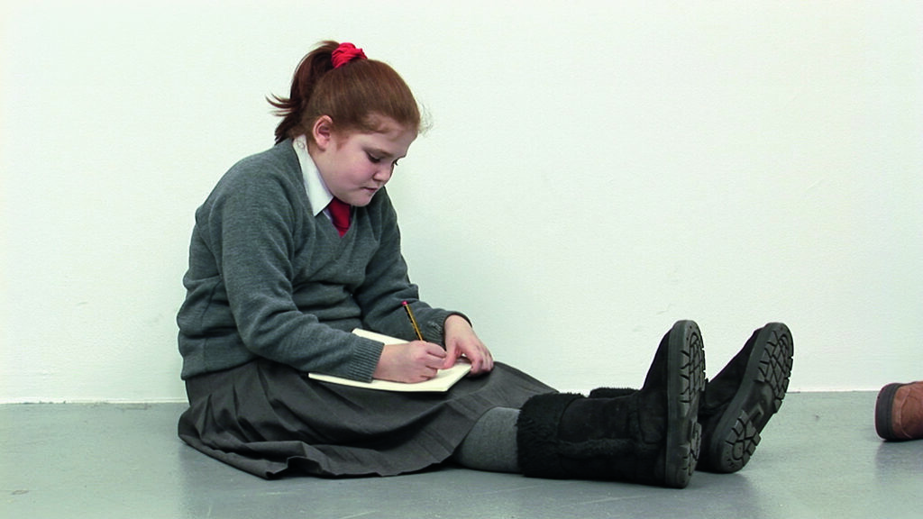 Video still of a young girl in a gray sweater sitting on a floor and drawing