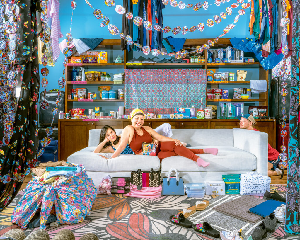 Two women seated on a messy couch in a cluttered room, surrounded by a disorganized assortment of items.