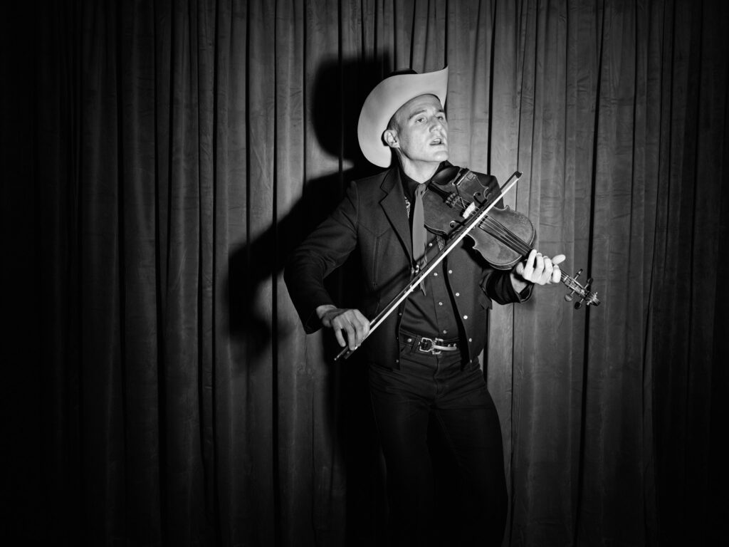 A black and white photo of a man donning Western wear playing the violin in front of a dramatic curtain.