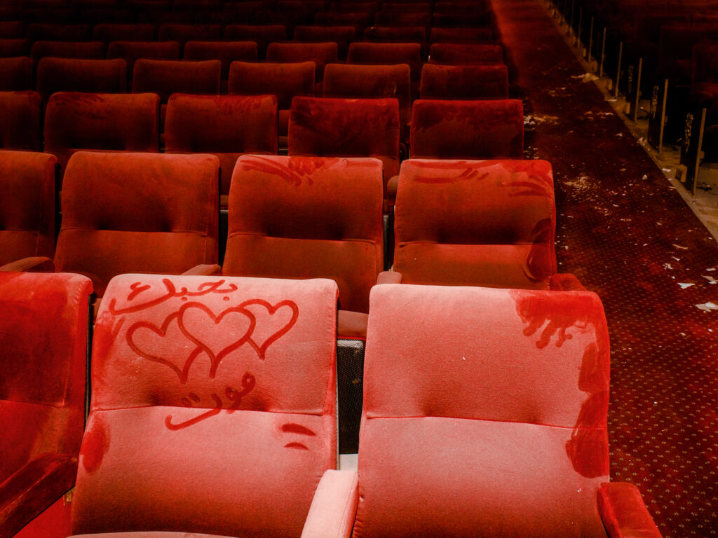 An empty theater with deep red velvet seats covered in dust. Arabic text and hearts are drawn into the layer of dust.