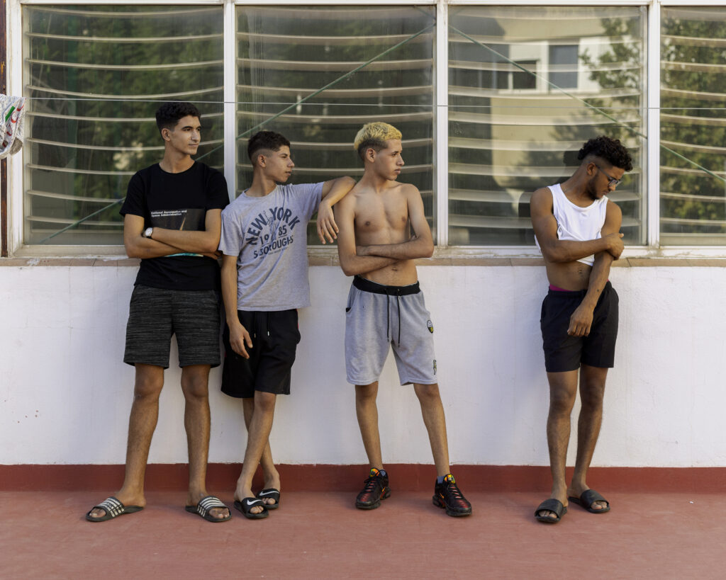 Four young men stand outside a nondescript building looking at each other.