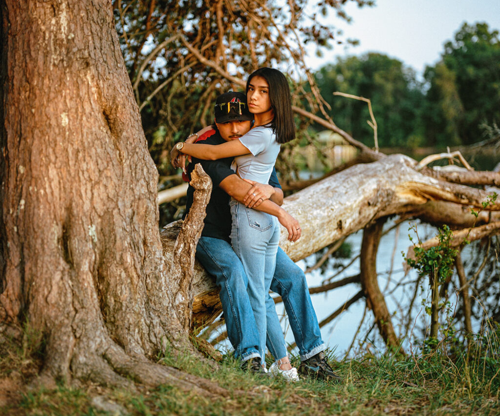 Two young individual—one female presenting, one male presenting—embrace while sitting on a fallen tree branch.