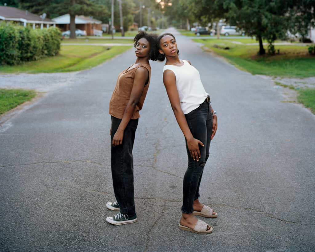 Two individuals lean head to head up against each other in the middle of a road.