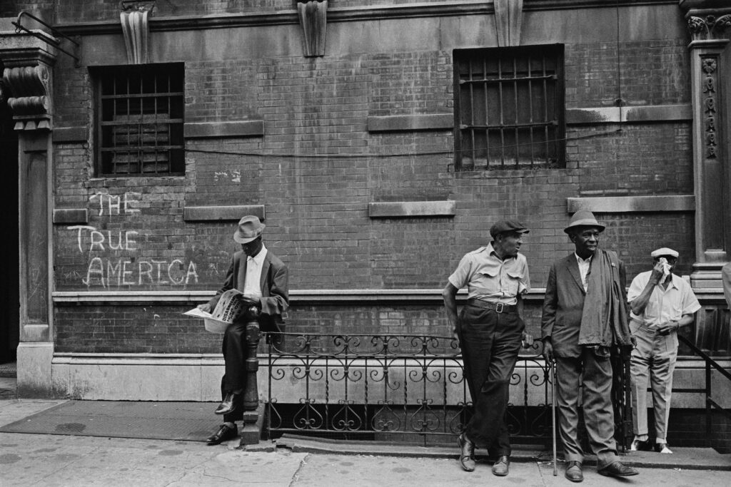 Four men in 60s-era clothing stand on a sidewalk. Written on the building behind them is "The True America."