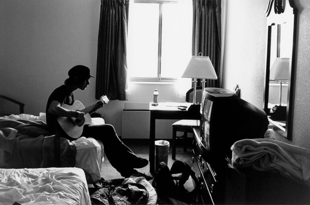 A young man sits at the edge of a bed in what appears to be a motel room playing guitar.
