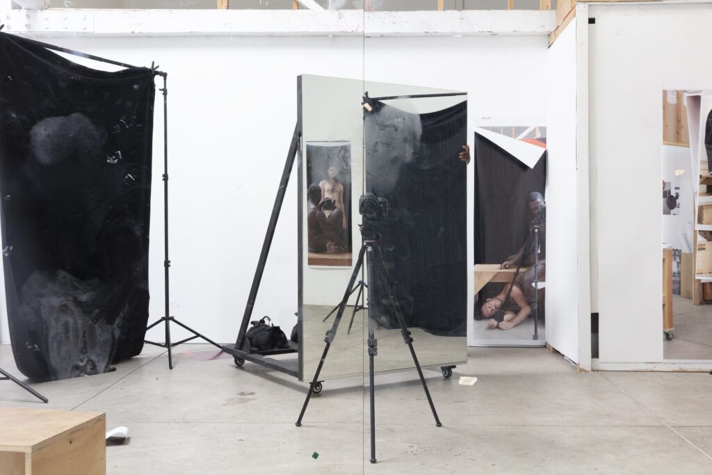 A tripod stands in the middle of a studio reflected in a mirror along with various individuals entwined around a camera.