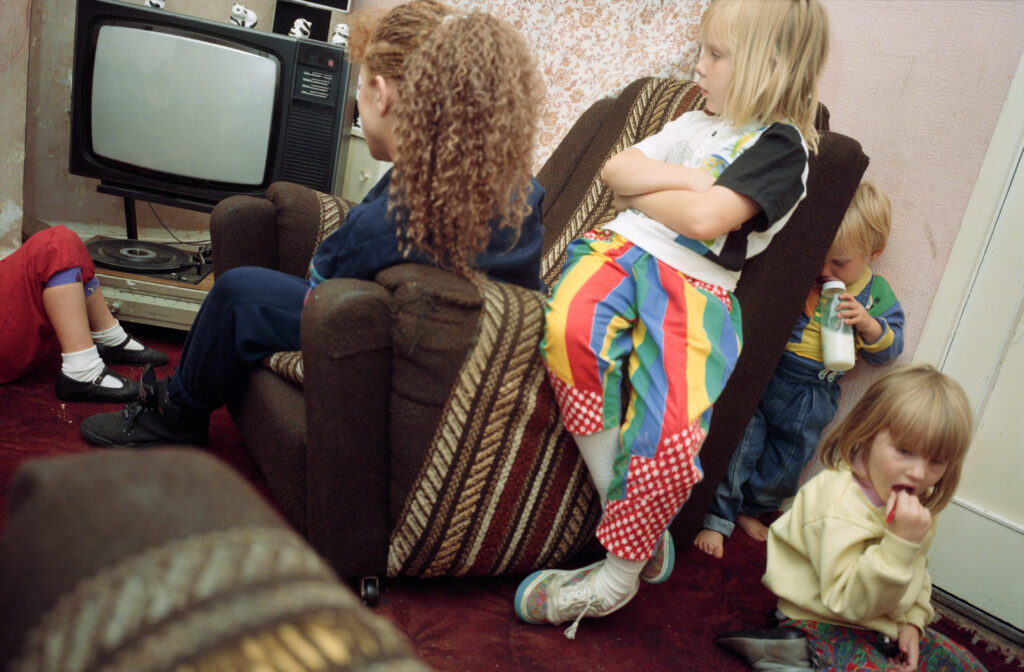 A small child leans against a chair occupied by an older woman. They look at a powered off TV.