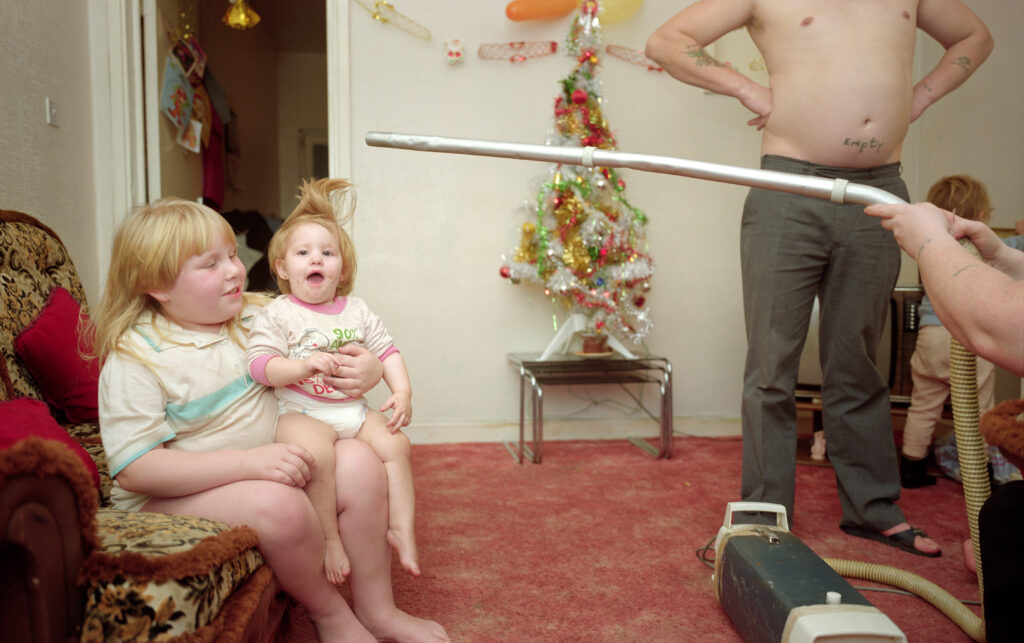 In a living room, an individual hold a vacuum extension above a toddler's head to make her hair stand straight up.