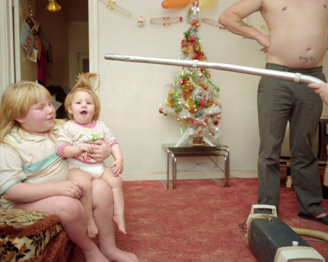 In a living room, an individual hold a vacuum extension above a toddler's head to make her hair stand straight up.