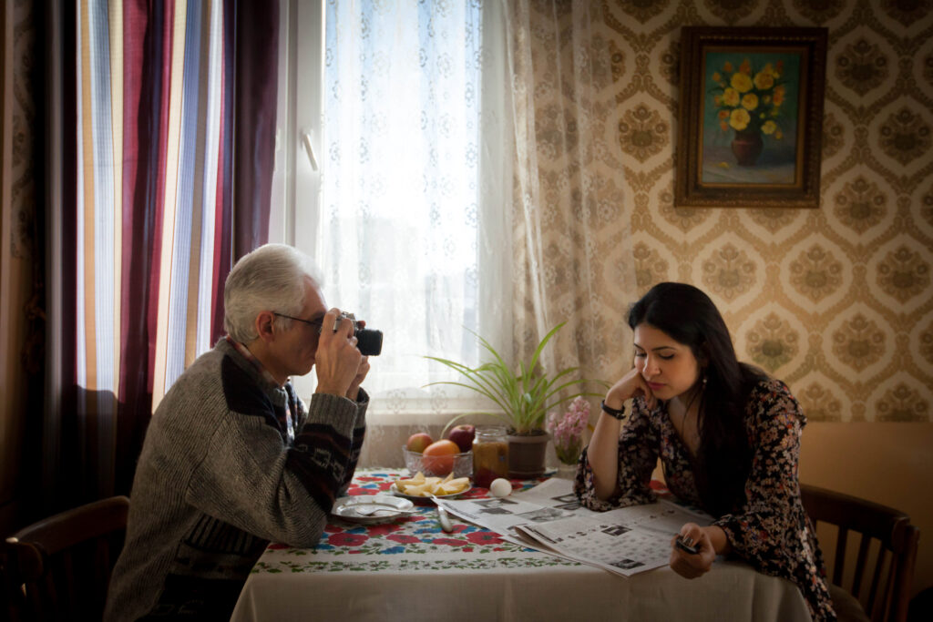 An older man sits at a table with his camera pointed at a younger woman sitting across from him at the table.