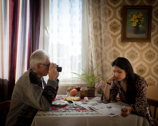 An older man sits at a table with his camera pointed at a younger woman sitting across from him at the table.