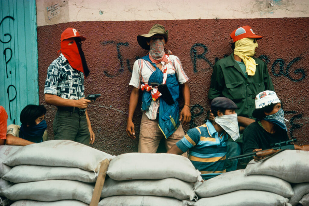 Six masked individuals stand and crouch behind a wall of sand bags. Some carry weapons.