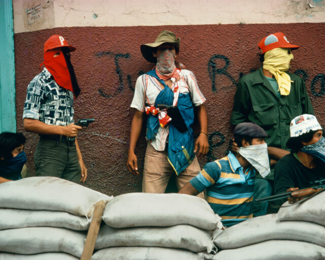 Three masked individuals stand behind a sand bag wall