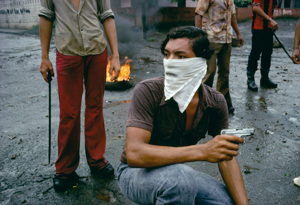 A man crouches on a street wearing a handkerchief and holding a small pistol.