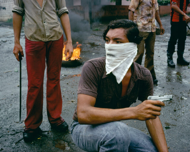 A man crouches on a street wearing a handkerchief and holding a small pistol.