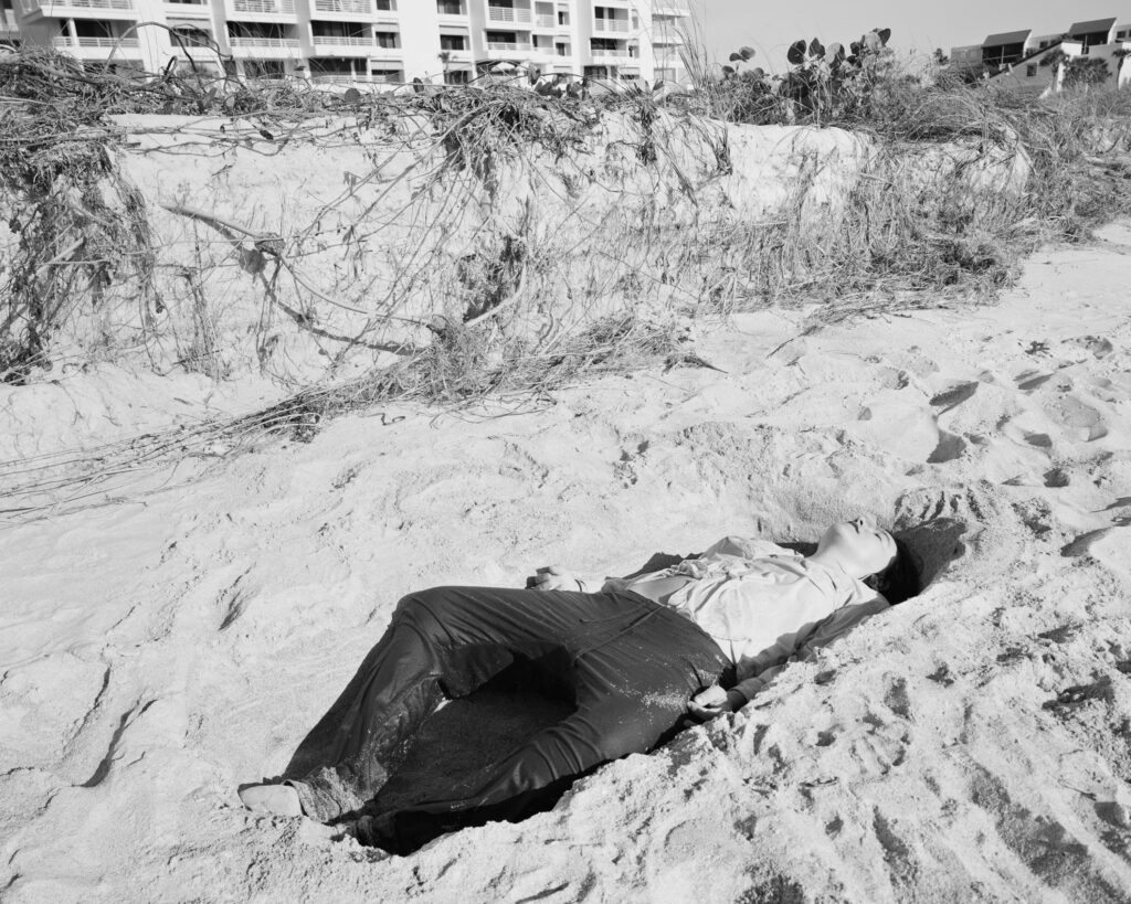 Alana Perino, Bay Isles Private Beach, 2020; from the series Pictures of Birds A black and white photo of an individual lying in a hole in the sand.
