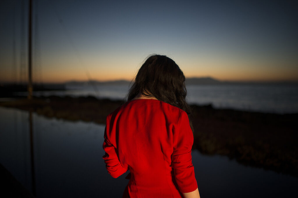Todd Hido, #11506-3940, 2016, from Intimate Distance (Aperture, 2025). © Todd Hido A woman in red looks out towards a dimming sunset.