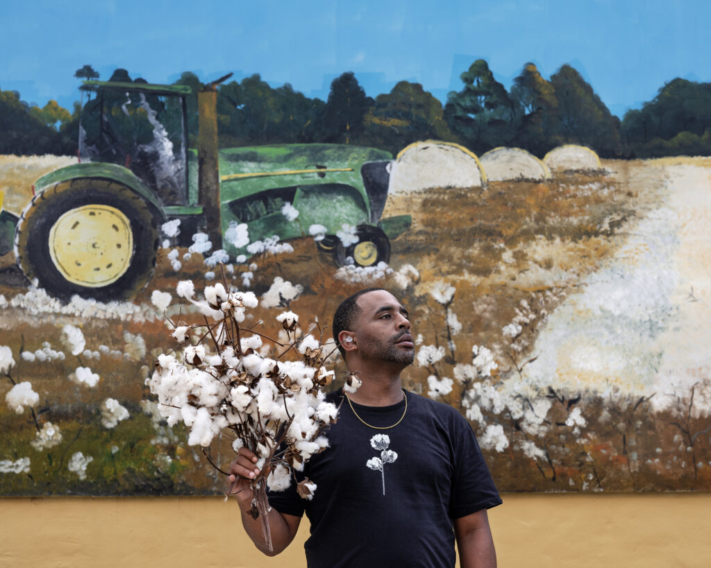 Fifth Generation Farmer, Garsburg, NC, 2024 A black man holding a bundle of cotton stands in front of a mural.
