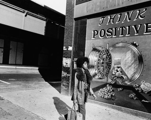 A black woman walks in front of a mural on the street that says