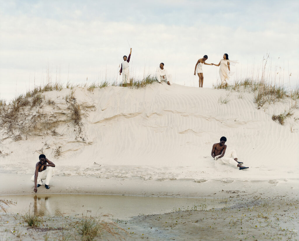 Black men and women dressed in white pose against amongst sand dunes.