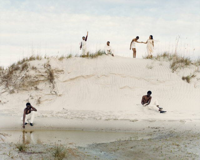 Black men and women dressed in white pose against amongst sand dunes.